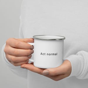 May include: White enamel mug with a silver rim, featuring the phrase "Act normal" in black text. The mug is held in a person's hands, showcasing its design and size. A simple, yet humorous, everyday item.