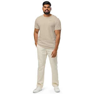May include: A beige short-sleeved crew neck t-shirt with a small black text logo on the left chest, paired with off-white trousers and white sneakers. The t-shirt features a simple design, a standard fit, and a smooth fabric texture throughout the garment.
