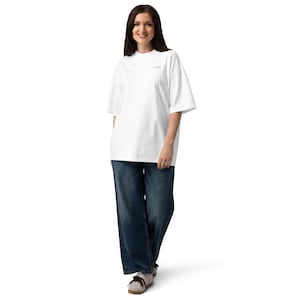 May include: A white oversized t-shirt with short sleeves and a small black text graphic on the chest, paired with loose-fitting dark blue denim jeans and light-colored sneakers. The t-shirt features a crew neckline and a relaxed, boxy fit.