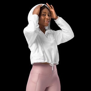 May include: A white windbreaker jacket featuring a hood, long sleeves, and a front zip closure. The jacket has a drawstring hem with toggles and a cropped length. It is paired with mauve leggings that feature side pockets on the thighs.