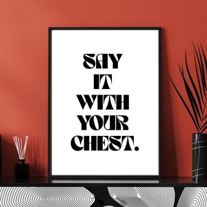 May include: Black framed art print with the text "SAY IT WITH YOUR CHEST" in bold, black font. The print is displayed on a black cabinet against a red wall. A black vase with a plant and a black diffuser are also visible.