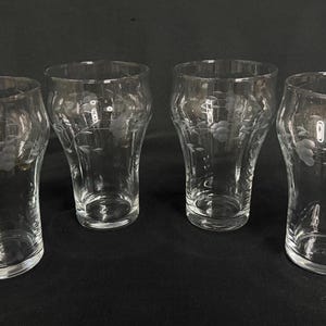 May include: Four clear, etched glass tumblers with a floral design. The glasses have a flared shape and are displayed against a dark background. Each glass features a delicate etched floral pattern.