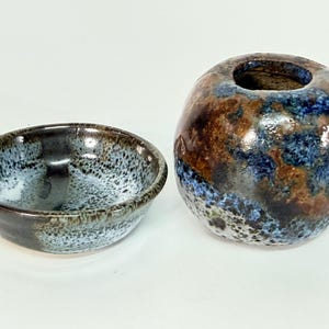 May include: A small ceramic bowl and a matching spherical vase featuring a mottled glaze in shades of deep blue, brown, and white. Both pieces have a glossy finish with a speckled texture, showcasing a blend of dark and light earthy tones.
