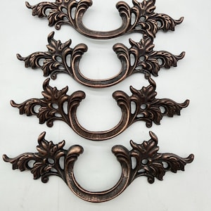 May include: Four bronze-colored drawer pulls with an ornate, floral design. Each pull has a curved handle and decorative flourishes on either side. The pulls are arranged in two rows of two against a white background, showcasing their intricate details.