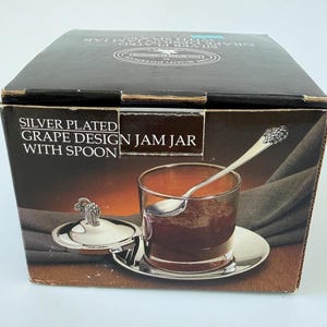 May include: Silver-plated jam jar with grape design, including a spoon and lid. The jar is filled with a dark red preserve and sits on a silver-colored plate. The product is packaged in a black box with text.
