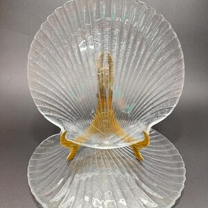 May include: Two clear glass seashell-shaped plates. The plates have a ribbed texture and are supported by a gold-colored metal stand. The plates are designed to resemble a scallop shell.