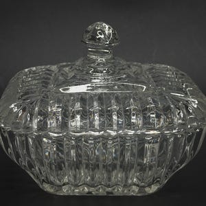 May include: A clear glass candy dish with a matching lid, featuring a square shape with rounded corners and a vertical ribbed pattern along the sides. The lid includes a small, faceted knob handle at the center, and the entire piece is made of transparent, pressed glass with a reflective surface.