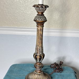 May include: A decorative table lamp with a bronze finish. The lamp features a detailed, ornate design with a fluted column and a decorative base. The lamp has a white bulb and a brown electrical cord.