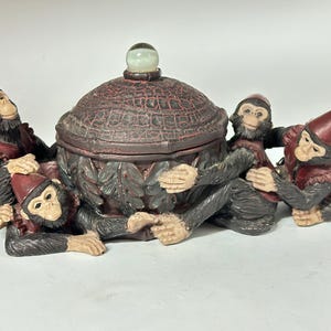 May include: A decorative trinket box featuring a central, textured, brown lid with a green glass orb on top. The box is surrounded by four monkey figurines wearing red fez hats and vests. The monkeys are in various poses, adding a playful touch.