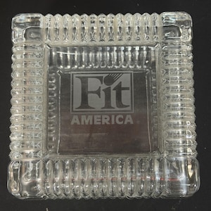 May include: A square clear glass ashtray featuring a thick, ribbed border and a smooth center panel. The center is etched with the text Fit AMERICA, with the letter i in Fit stylized to resemble a tennis racket or similar sports equipment.