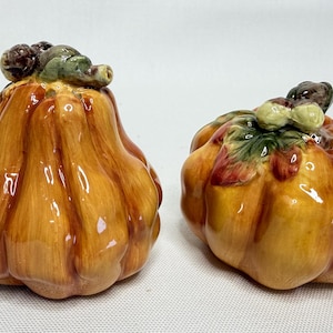 May include: Two ceramic pumpkin figurines. The pumpkins are orange with brown and green accents on top. The pumpkins have a glossy finish and are likely decorative items for the fall season.