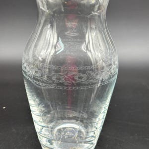 May include: A clear glass vase with a gold rim and a decorative etched floral pattern around the middle. The vase has a flared top and a slightly curved body. The base is clear and the vase is empty.