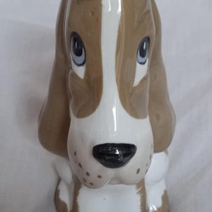 Könnte beinhalten: Keramikfigur eines Basset Hound-Hundes. Der Hund hat braune und weiße Markierungen mit einem weißen Streifen auf dem Kopf. Er hat blaue Augen und eine schwarze Nase. Die Figur sitzt aufrecht.