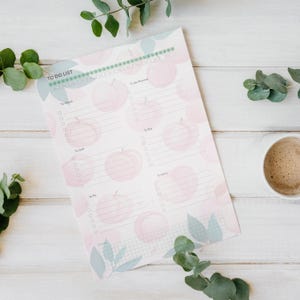 May include: A pastel-colored To Do List notepad with a peach pattern. The notepad has a spiral binding and sections for tasks like "To Check," "To Call," and "To Buy." Green leaves and a cup of coffee are in the background.