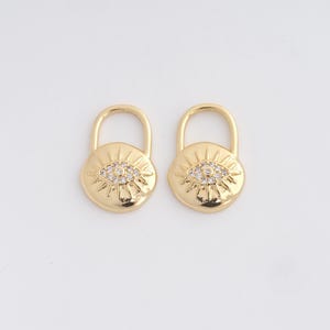 May include: Two gold-colored padlock charms. Each charm features a sunburst design with a central eye motif encrusted with small, clear crystals. The padlocks have a rounded shape and a curved handle.