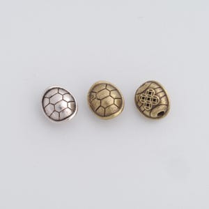 May include: Three metal turtle-shaped beads in silver, gold, and bronze. Each bead has a textured, segmented pattern resembling a turtle shell. The bronze bead features a decorative four-leaf clover design.