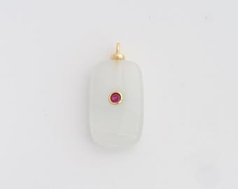 Puede incluir: Un colgante rectangular con una argolla dorada y una pequeña gema roja y redonda engastada en el centro. El colgante es de color claro y opaco, sobre un fondo blanco.