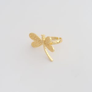 May include: A gold-coloured ring with a dragonfly design. The dragonfly has detailed wings and a slender body. The ring is adjustable and sits on a white background. This jewellery item is suitable for everyday wear.