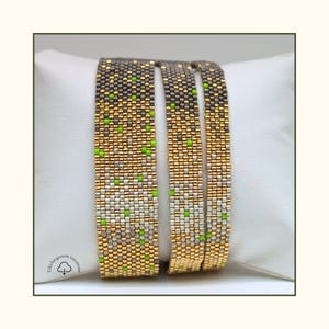 Peut inclure: Trois bracelets perlés avec un motif dégradé. Les bracelets présentent des perles dorées, argentées, grises et vertes. Les bracelets sont présentés sur un oreiller blanc. Les bracelets sont faits de petites perles carrées.
