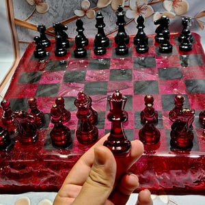 Könnte beinhalten: Ein handgefertigtes Schachspiel mit einem rot-schwarz karierten Brett. Die Schachfiguren sind schwarz und rot, mit einer Königinfigur im Vordergrund. Das Brett hat eine glänzende Oberfläche, und das Set wird auf einer hellen Oberfläche präsentiert.