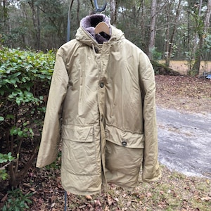 Puede incluir: Un abrigo de invierno color caqui con capucha y cuello forrado de piel cuelga al aire libre. El abrigo tiene dos bolsillos delanteros y cierre de botón. El abrigo está colgado de un gancho de metal frente a un fondo de árboles y follaje.