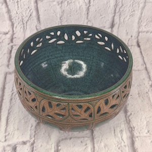 May include: A round ceramic bowl featuring a deep teal interior glaze and a brown exterior with a cut-out leaf pattern. The rim is also finished with a teal glaze, and the cut-out design creates a series of open leaf shapes around the upper section.