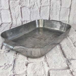 May include: A rectangular silver-coloured metal baking pan with a smooth interior surface and rounded corners. The pan features two integrated handles on opposite ends, each detailed with three horizontal embossed lines for a textured grip.