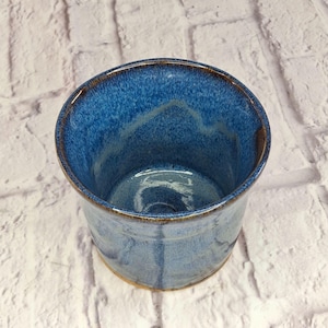 May include: A cylindrical ceramic mug featuring a deep blue glaze with mottled lighter blue and white accents. The interior and exterior surfaces display a glossy finish with subtle variations in color depth and a smooth, rounded rim edge.