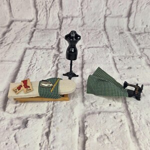 Puede incluir: Un set de costura en miniatura que incluye un maniquí negro, una tabla de planchar de madera con funda de cuadros verdes y blancos, una plancha de metal pequeña, una cinta métrica amarilla, dos etiquetas rectangulares y una máquina de coser.