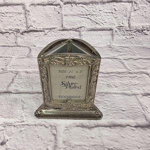 Może przedstawiać: Posrebrzana ramka na zdjęcia z ozdobnymi detalami. Ramka ma wymiary 6,35 cm x 7,62 cm i zawiera napis "Silver Plated" i "Godinger". Ramka ma dekoracyjne, łukowate zwieńczenie i prostokątną podstawę.