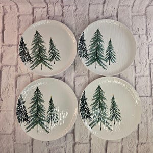Puede incluir: Un juego de cuatro platos redondos de cerámica blanca, cada uno con una superficie texturizada y un diseño impreso de tres árboles de hoja perenne en tonos verde oscuro y negro, con una pequeña estrella dorada en la punta del árbol más alto.