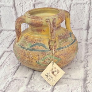May include: A round ceramic vase with two handles, featuring a mottled yellow and brown finish with green geometric patterns. A beige paper tag is attached to one handle, printed with the text River Junction Pottery Works in black ink.