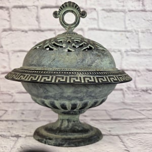 Può includere: Un bruciatore di incenso in stile antico, color verderame. Il vaso con coperchio presenta un motivo traforato decorativo sulla parte superiore e un motivo a chiave greca attorno al bordo. Ha una base a piedistallo e un manico con un'apertura circolare.