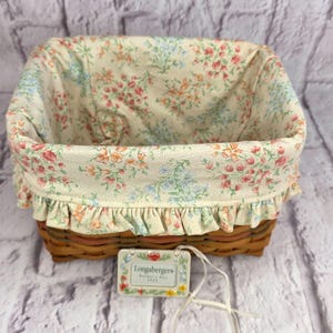 May include: A rectangular woven brown basket featuring a fabric liner with a cream background and a floral pattern in pink, orange, and blue. The liner has a ruffled edge and a rectangular tag attached that reads Longaberger Mother's Day 2003.