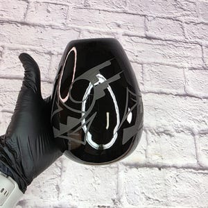 May include: A black glass vase with a unique shape and a gray abstract design. The vase is held by a gloved hand against a white brick wall background. The vase has a glossy finish.