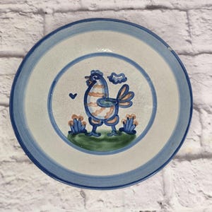 Könnte beinhalten: Ein runder Keramikteller mit handgemaltem Design. Der Teller hat einen blau-weißen Rand und eine zentrale Illustration eines Vogels mit blauen, orangefarbenen und weißen Streifen. Der Vogel steht auf einer grünen Basis mit orangefarbenen und blauen floralen Akzenten.