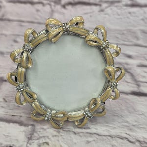 May include: A round picture frame with a silver-colored rim and a series of cream-colored bows around the edge. Each bow is accented with small, clear crystals. The frame is designed to hold a circular photo.