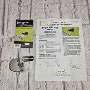 Puede incluir: Un accesorio de metal plateado para doblar bies en máquinas de coser, acompañado de una hoja de instrucciones impresa y una tarjeta de empaque. El texto en los materiales especifica un ancho de 28 mm y compatibilidad con modelos Baby Lock.