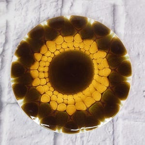 Könnte beinhalten: Ein runder, dekorativer Untersetzer mit einem einzigartigen Design. Der Untersetzer hat einen dunkelbraunen Mittelkreis, umgeben von gelben und olivgrünen Formen, die ein Blumen- oder abstraktes Muster bilden. Mit weißem Rand. Durchmesser ca. 10 cm.