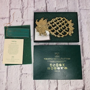 May include: A gold-toned metal pineapple trivet by Virginia Metalcrafters, presented in a dark green box. The set includes a descriptive paper insert, a small tag, and a green felt pad, all featuring the Virginia Metalcrafters brand name.