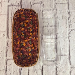May include: An oval basket with a fabric lining featuring autumn leaves in red, orange, and yellow. Two clear plastic containers are next to the basket. The basket has a woven, light brown border.
