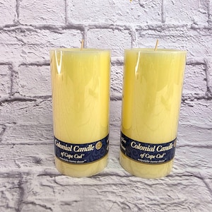 May include: Two yellow pillar candles with wicks, labeled "Colonial Candle of Cape Cod." The candles are cylindrical and wrapped in clear plastic. The background is a white brick wall.