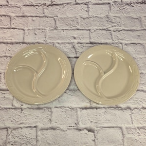 Puede incluir: Dos platos divididos de color crema. Cada plato tiene un divisor curvo que crea tres secciones. Los platos tienen un diseño simple y elegante con un borde ligeramente elevado. Los platos son de cerámica.