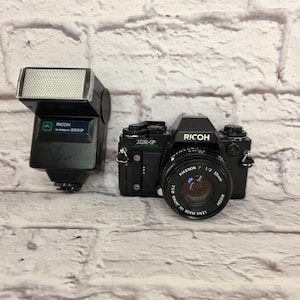 Peut inclure: Un appareil photo argentique 35 mm Ricoh XR-P noir avec un objectif Rikenon P 1:2 50 mm et un flash Ricoh XR Speedlite 300P noir séparé. Le boîtier de l'appareil porte le logo Ricoh, le nom du modèle et divers cadrans, tandis que le flash a un panneau avant blanc texturé.