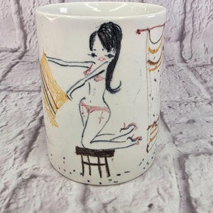 Può includere: Una tazza in ceramica bianca con un'illustrazione a schizzo in nero e rosa di una figura dai capelli scuri inginocchiata su un piccolo sgabello. La figura tiene in mano un pezzo di tessuto giallo e un drappeggio è visibile sul lato destro.