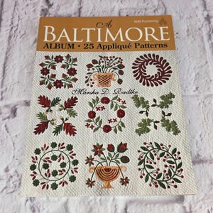 May include: A softcover book titled A Baltimore Album: 25 Appliqué Patterns by Marsha D. Radtke. The cover features a grid of nine distinct floral and botanical appliqué designs in shades of red, green, and tan on a textured white background.