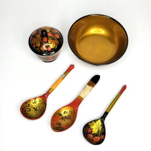 May include: A collection of Russian nesting dolls, a bowl, and wooden spoons. The items are painted with floral designs in red, gold, and black. The bowl has a gold interior. The spoons have a red handle and a gold bowl.