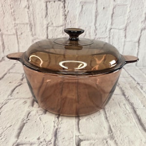 Peut inclure: Une cocotte ronde en verre brun translucide dotée de deux poignées latérales intégrées et d'un couvercle en verre assorti avec un bouton central. L'ustensile de cuisine présente une surface lisse et polie ainsi qu'un récipient profond.