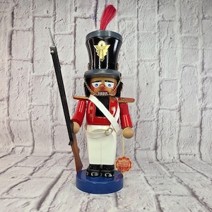 May include: A wooden nutcracker soldier figure wearing a black hat with a red feather, a red jacket with gold accents, and white trousers. The figure holds a tall black rifle and stands on a round blue base, with a small red tag attached to its arm.