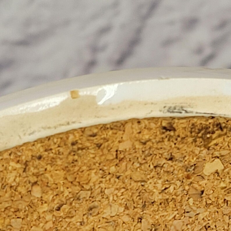 May include: Close-up of a white ceramic coaster with a cork base. The coaster has a slightly uneven edge and a visible chip. The cork base is made of small, irregular pieces.